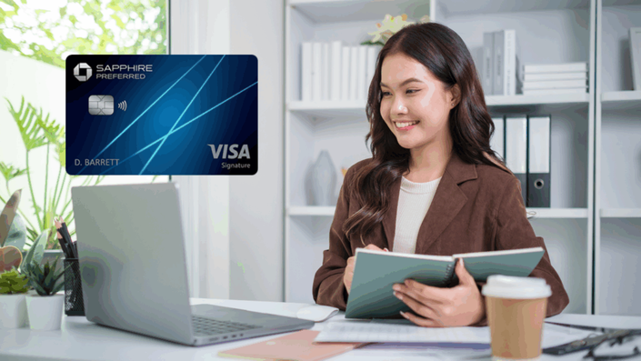 A woman sitting at a desk, smiling and holding a notebook with a Sapphire Preferred Visa Signature card visible in the foreground. A coffee cup and laptop are on the desk, and the background features bookshelves with plants.