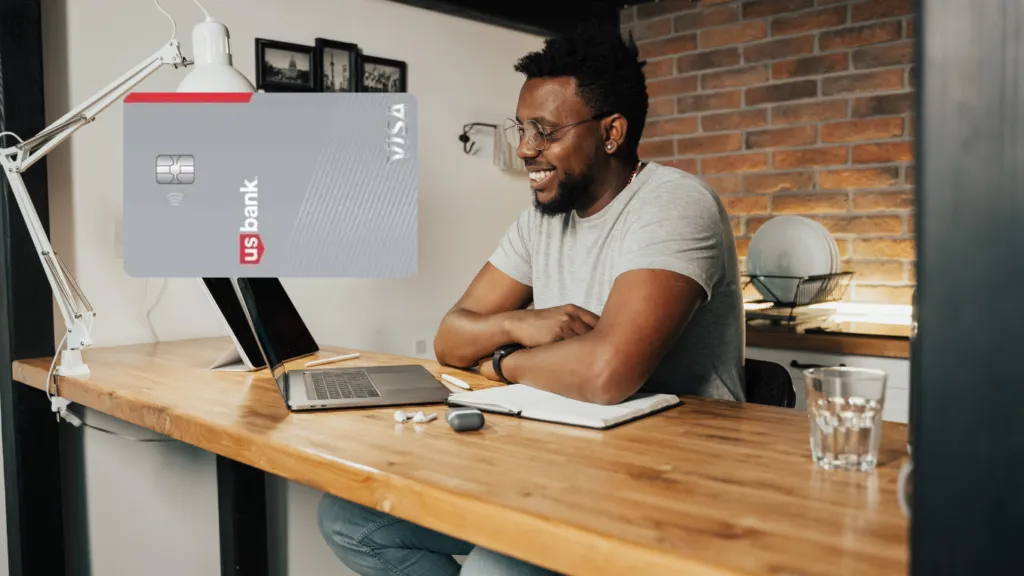 Un hombre con barba, gafas y una camiseta gris sonríe mientras trabaja en su laptop en un escritorio de madera con un fondo de pared de ladrillos