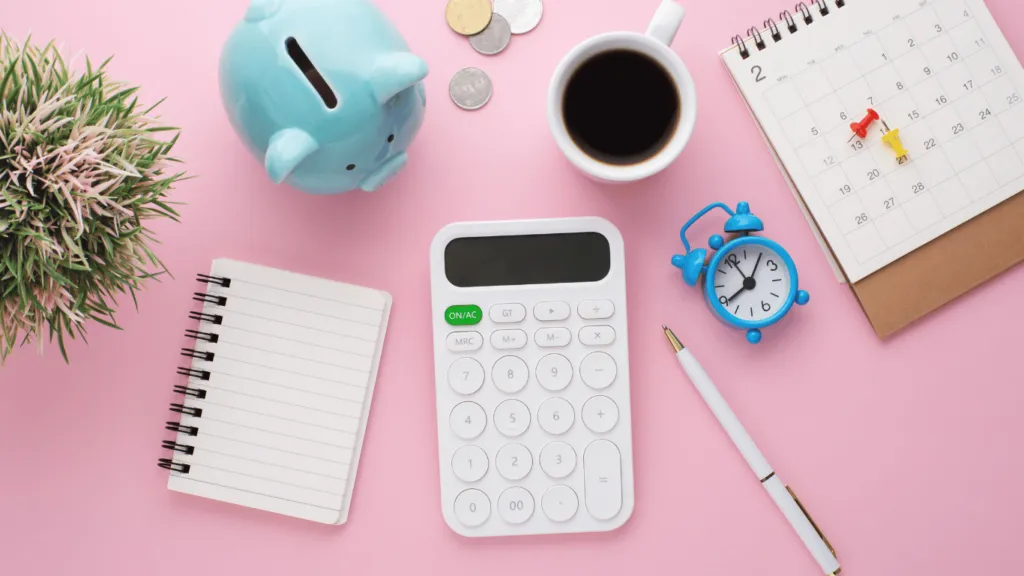 Vista superior de una mesa rosa con una alcancía azul en forma de cerdito, una calculadora blanca, una taza de café negro, un cuaderno en espiral con hojas en blanco, un bolígrafo, un pequeño reloj despertador azul, monedas sueltas, un calendario con chinchetas de colores y una planta decorativa.