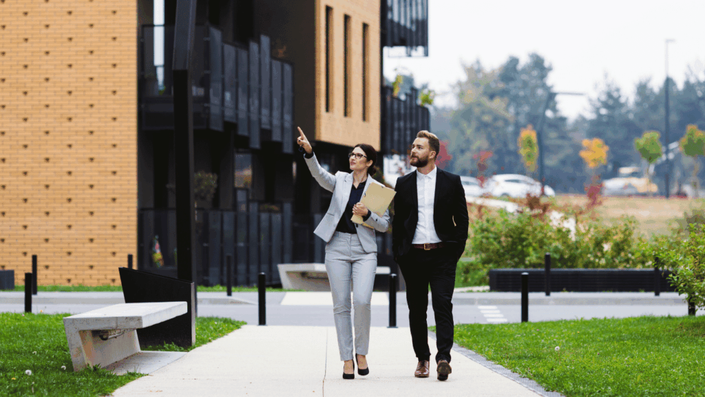 Agente mostrando propiedades de bienes raíces a un cliente en Estados Unidos, caminando juntos frente a un edificio moderno durante una visita inmobiliaria.