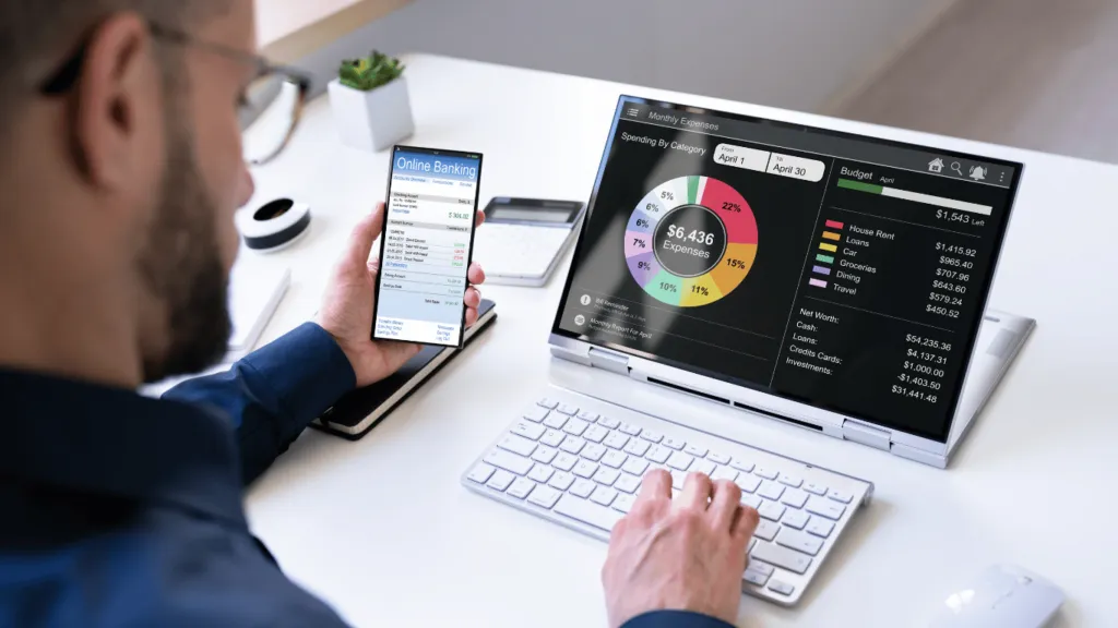 A person using a budgeting app on their laptop and smartphone, tracking expenses and setting financial goals for better money management.