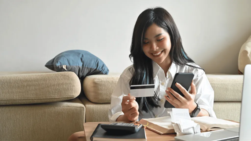 A young woman sitting on a couch, smiling while holding a smartphone and a credit card, seemingly making an online purchase. She appears to be managing her finances and looking for the best credit card for online shopping.