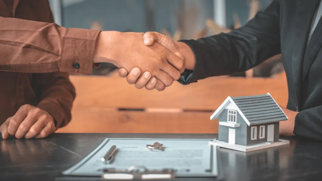 Two individuals shaking hands over a real estate contract with a house model in the background, symbolizing a successful deal in investing in real estate.