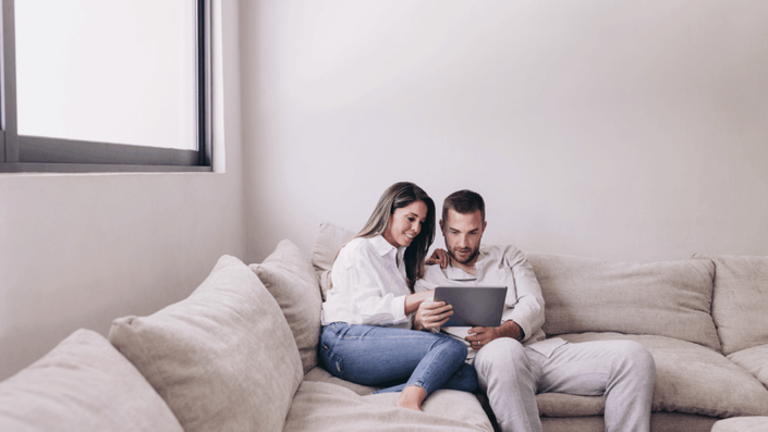 Pareja joven sentada en un sofá claro, mirando juntos una tablet y sonriendo, en un ambiente hogareño y relajado, simbolizando la planificación financiera en pareja.