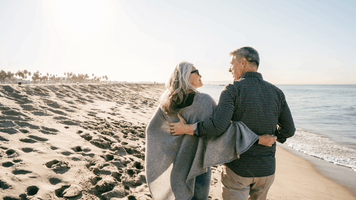 Pareja de personas mayores caminando abrazados por la playa al atardecer, disfrutando de su jubilación y de un momento tranquilo junto al mar.