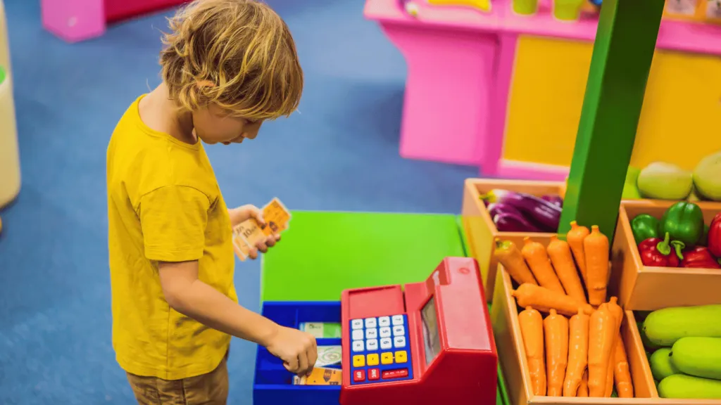 Niño jugando en una tienda de juguete con una caja registradora y billetes de juguete. Actividad lúdica sobre Educación Financiera para Niños.