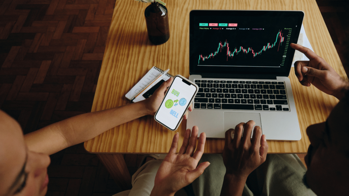Dos personas observan gráficos en una computadora portátil y un teléfono móvil. En la pantalla del teléfono, se ven los íconos de "Comprar" y "Vender" relacionados con inversiones, mientras analizan datos financieros en el portátil.