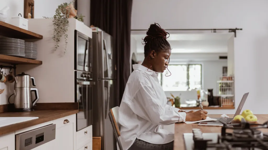 A determined freelancer working from home, taking notes while using a laptop. A great example of how to start a business with little money in a home-based setup.