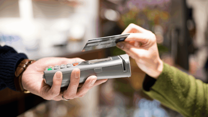 A close-up of a person holding a credit card above a payment terminal for a Tap-to-pay transaction, with another person holding the card reader.