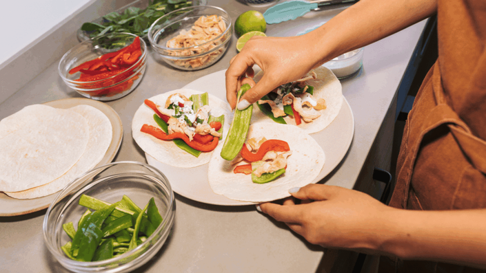 Manos preparando tacos en una cocina, con tortillas, pimientos, pollo y hierbas frescas sobre la mesa, mostrando un proceso de planificación de comidas económica y saludable.