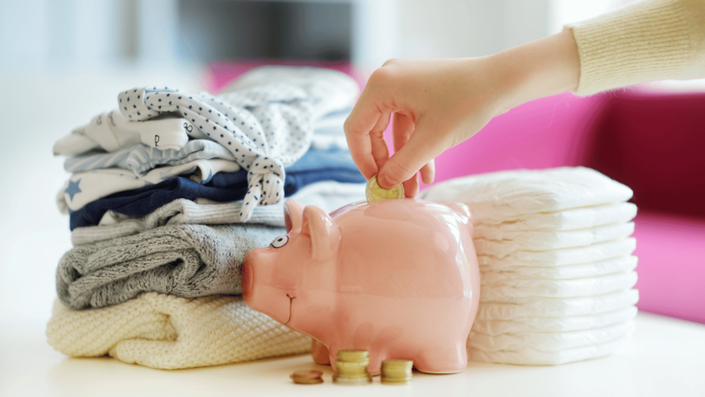 Persona ahorrando dinero en una alcancía junto a ropa y pañales de bebé, simbolizando la importancia de tener un presupuesto para un bebé a camino desde el inicio.