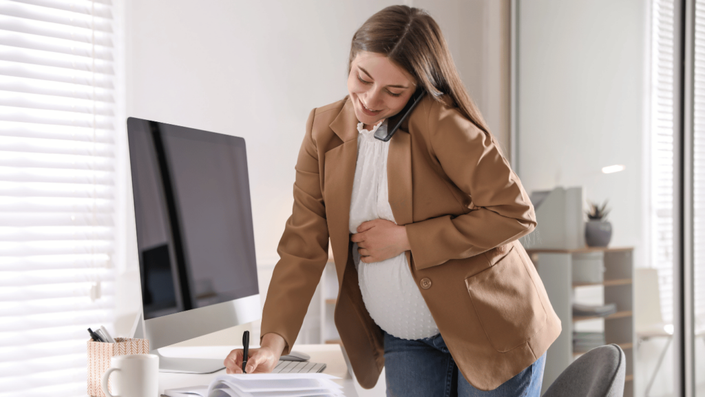 Mujer embarazada trabajando en la oficina mientras habla por teléfono y toma notas, representando la planificación de un presupuesto para un bebé a camino y la organización necesaria durante la licencia parental.