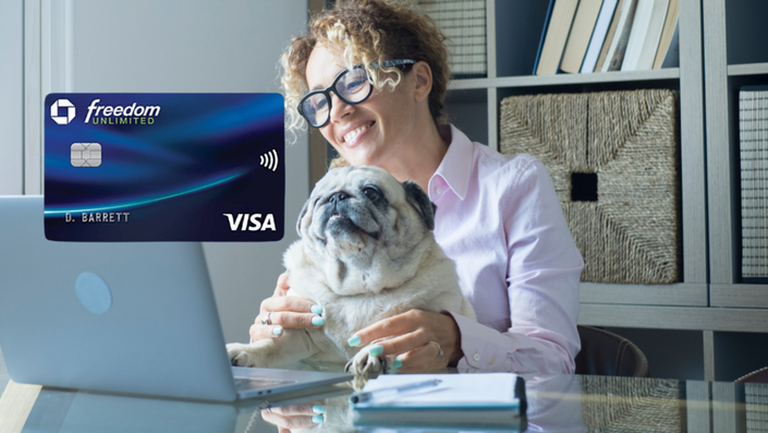 Una mujer de cabello rizado y oscuro, usando gafas y una camisa rosa, sonríe mientras sostiene a su perro pug en su regazo frente a una computadora portátil. En la pantalla de la computadora se ve una tarjeta de crédito Chase Freedom Unlimited con el logo de Visa.