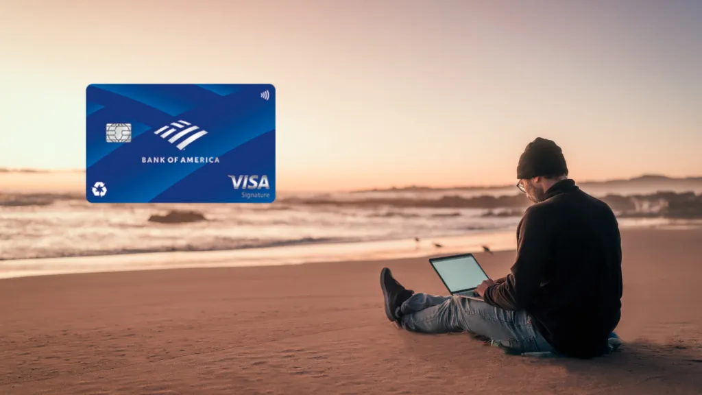 Hombre con gorro y gafas, sentado en la arena de una playa al atardecer, usando su laptop. Al fondo, el mar y el cielo con tonos cálidos.